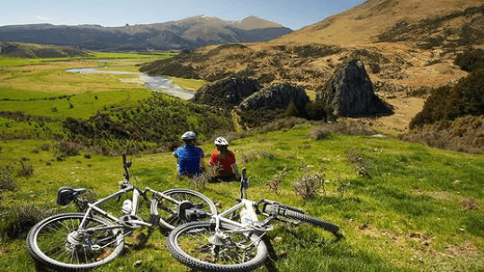 Two cyclists resting with their bikes on a scenic New Zealand cycle trail overlooking a river valley